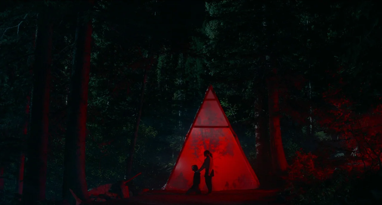 Trim Season (2023) movie still: wide — A dense coniferous forest is engulfed in darkness, punctuated only by a glowing structure. Two human…