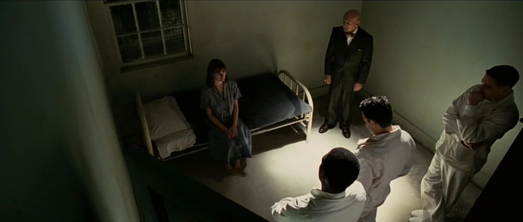 Shutter Island (2010) movie still: wide — A stark room with pale walls and a single barred window holds a metal cot. One woman sits on the bed while…