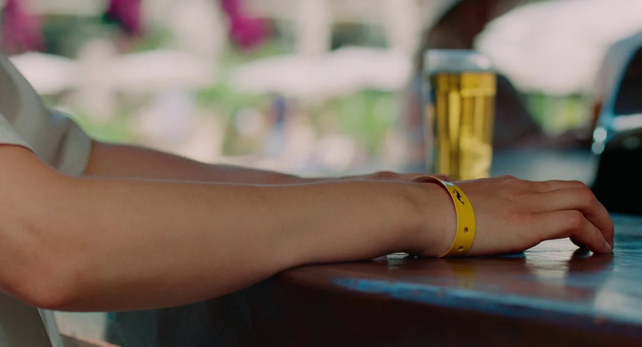 Aftersun (2022) movie still: closeup — An outdoor bar table surface holds a glass of beer in the blurred background. A person rests their arm and…