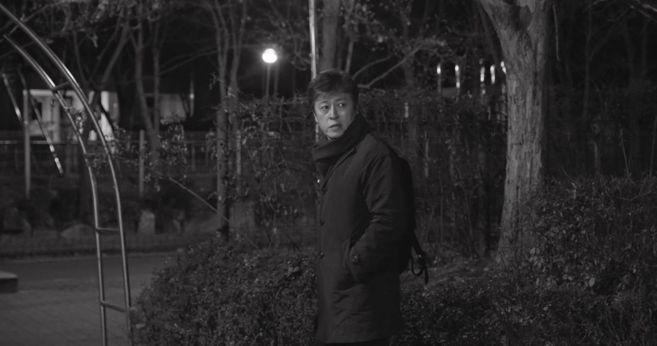 The Day After movie still: medium — A night park scene features bare trees and a metal arched structure in background. A middle-aged man in a dark…