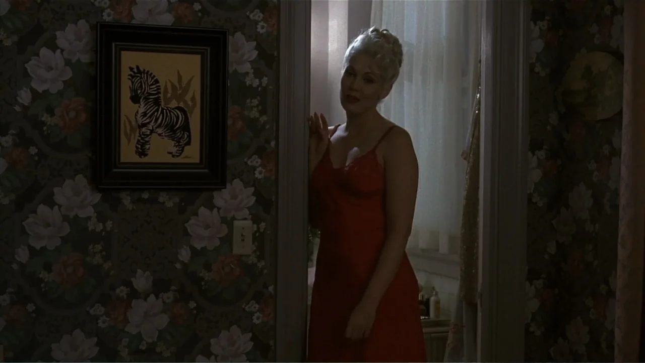 The Mambo Kings (1992) movie still: medium — A dimly lit bedroom doorway features floral wallpaper and a framed zebra print picture. A blonde woman…