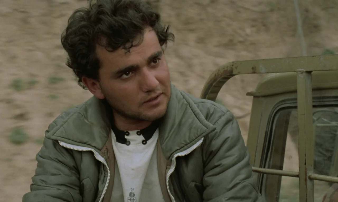 Through The Olive Trees (1994) movie still: medium — A man sits in the passenger seat of an open military-style vehicle. He gazes off-screen into…