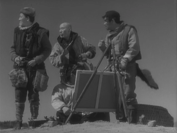 Scandal (1950) movie still: fullbody — Four men in heavy winter clothing gather outdoors on a sandy, barren landscape. One man sits by a wooden…