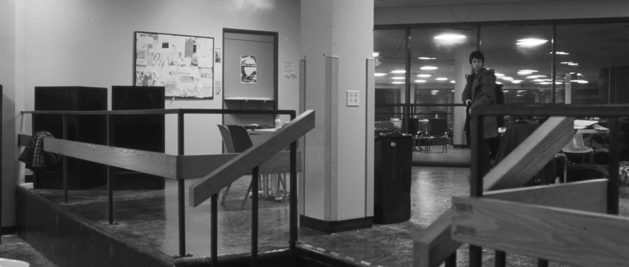 Polytechnique (2009) movie still: wide — A spacious room with wooden railings and a large glass-enclosed area beyond. A man stands alone holding a…