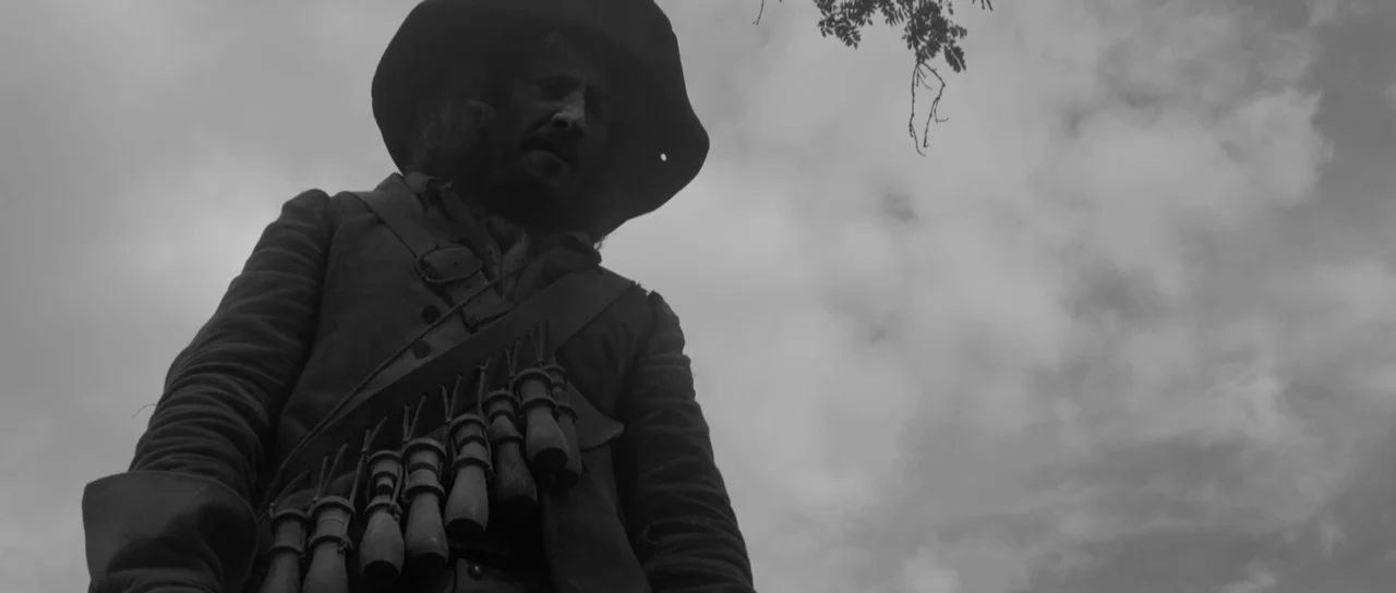 A Field In England (2013) movie still: medium — An overcast sky hangs above a field with a man in period clothing. The man wears a wide-brimmed hat…