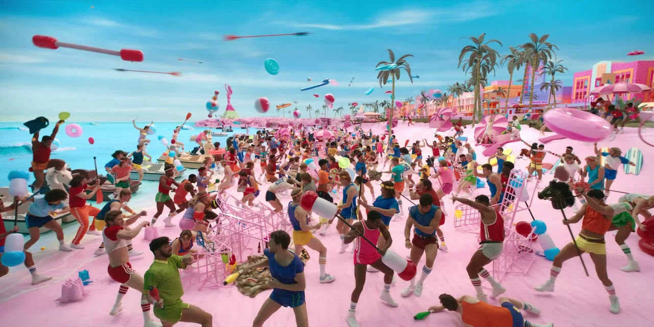 Barbie (2023) movie still: wide — A surreal beach with solid pink sand and bright blue ocean under a clear sky. A large crowd of people in colorful…