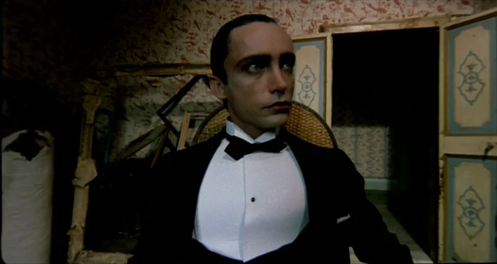 Blood For Dracula (1974) movie still: medium — A dimly lit room features patterned wallpaper and an open door frame. A man in formal evening wear…