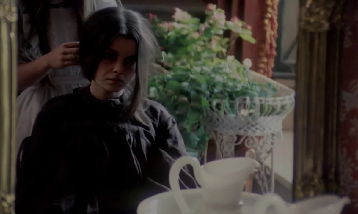 Kamouraska (1973) movie still: medium — A woman sits before a mirror in a room with a decorative potted plant. A child stands behind her and styles…