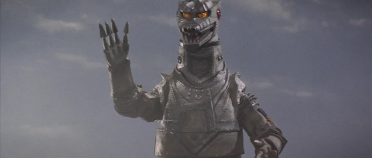 Terror of Mechagodzilla (1975) movie still: medium — A robotic monster stands against a flat grey overcast sky. The mechanical creature holds its…