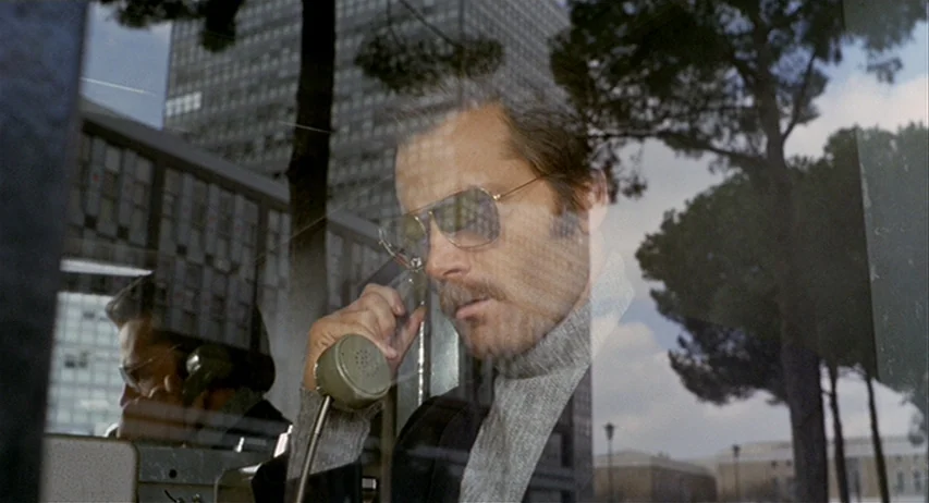 The Fifth Cord (1971) movie still: closeup — A man stands inside a glass telephone booth located on a paved outdoor path. The man holds a telephone…