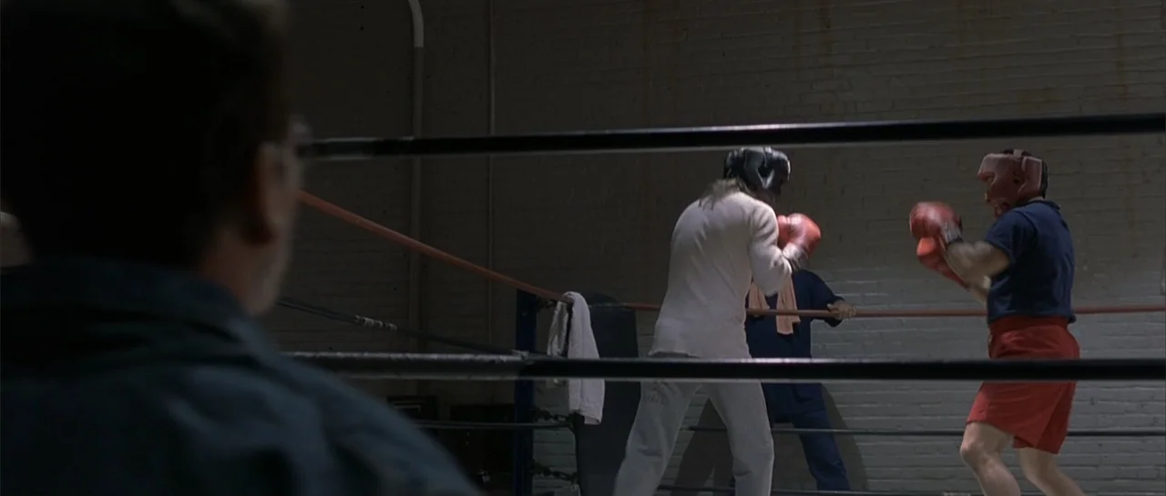 Bandits (2001) movie still: medium — A dimly lit indoor training area contains a boxing ring with dark ropes. Two men in headgear exchange punches…