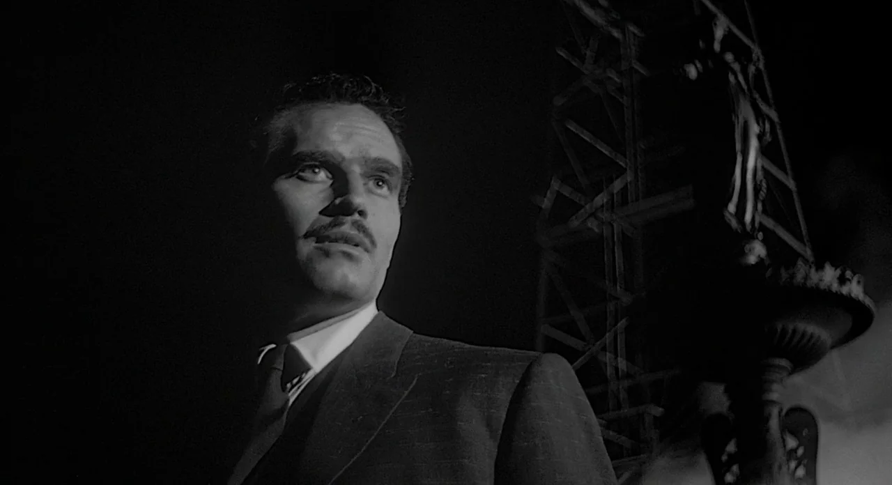 Touch of Evil (1958) movie still: medium — An industrial night setting features a large metal derrick looming in the dark background. A man stands…