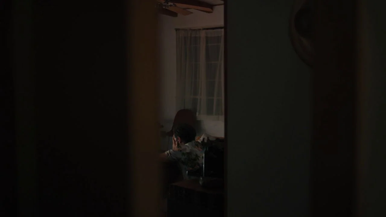 She Dies Tomorrow (2020) movie still: wide — A dark, dimly lit room contains a person seen through a narrow doorway. The person sits alone by a…