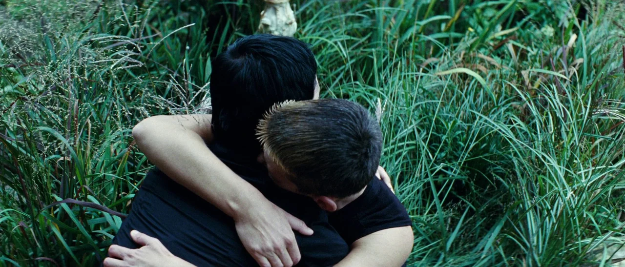 Outside Satan (2011) movie still: medium — Two individuals sit embraced amidst thick, overgrown green grass in a field setting. The figures are seen…
