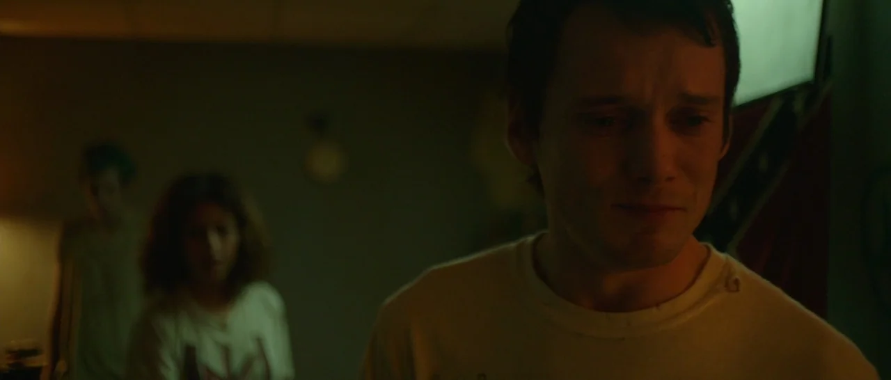 Green Room (2015) movie still: medium — A dimly lit basement features a concrete wall and blurred figures in background. A young man stands in the…