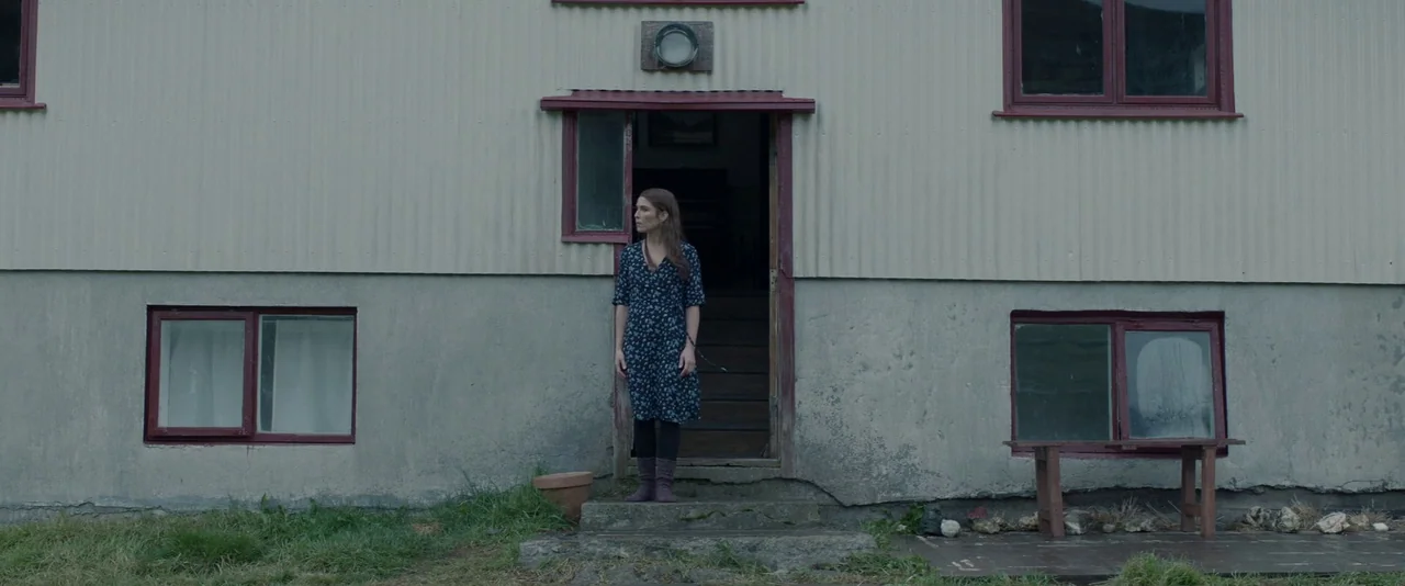 Babel (2006) movie still: fullbody — A house exterior features corrugated light-colored metal cladding and maroon-framed windows. A woman stands in…