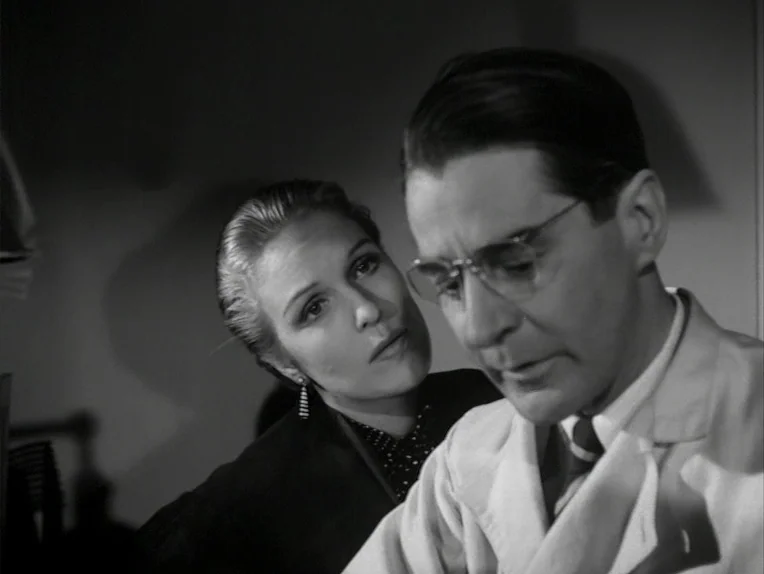 A Lesson In Love (1954) movie still: medium — A dimly lit room with high-contrast shadows defining the subjects' features. A woman stands behind a…