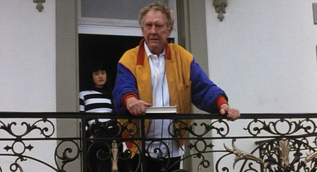 King Lear (1987) movie still: medium — A wrought-iron balcony overlooks a building exterior with white stucco walls. An elderly man stands in the…