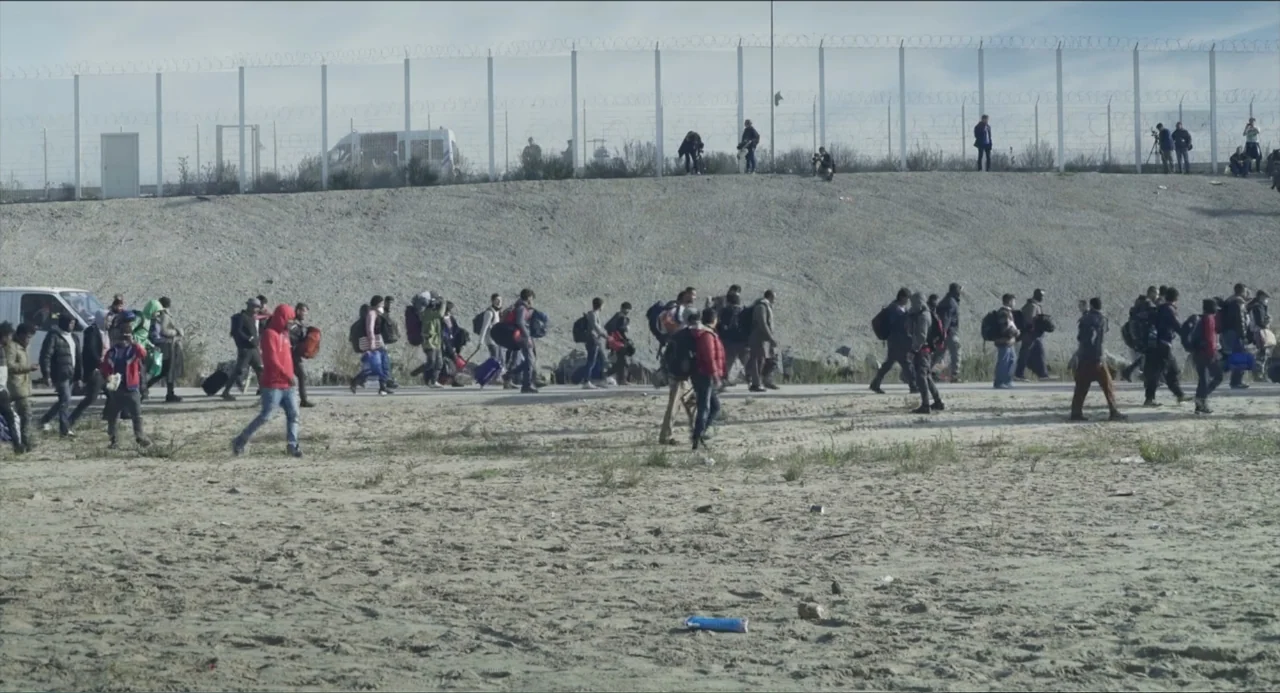 Human Flow (2017) movie still: wide — A wide expanse of sandy ground features a chain link fence along a ridge. A large group of people carrying…