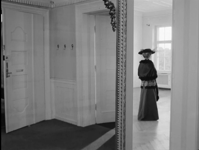 Effi Briest (1974) movie still: wide — A white-walled hallway features a tall ornate mirror reflecting a woman in period clothing. The woman stands…