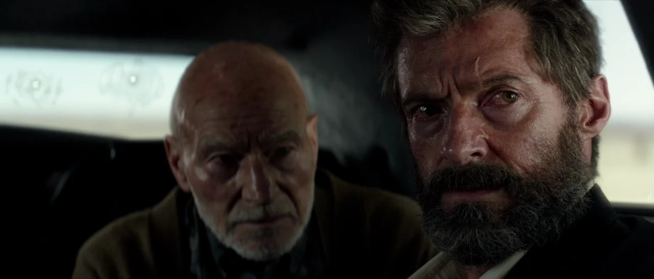 Logan (2017) movie still: medium — The interior of a car features dark upholstery and sunlight entering through the window. Two older men sit…