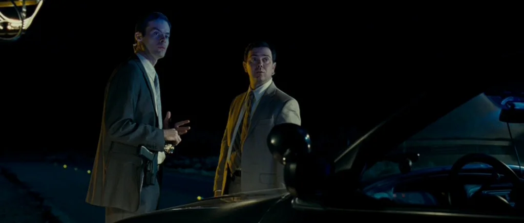 Paul (2011) movie still: medium — Two men in suits stand next to a dark sedan on a desolate road at night. One man gesture with his hand while they…