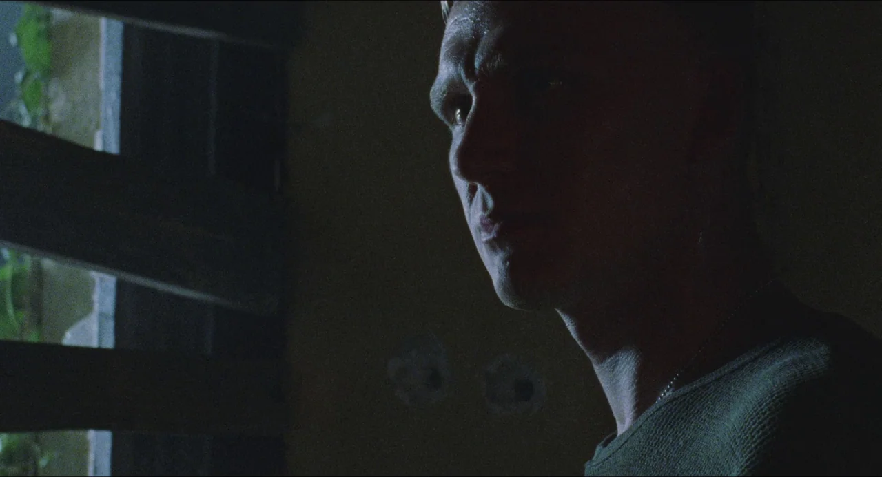 Dog Soldiers (2002) movie still: closeup — A dark indoor space with light filtering through horizontal slats of a window. A man stands in partial…