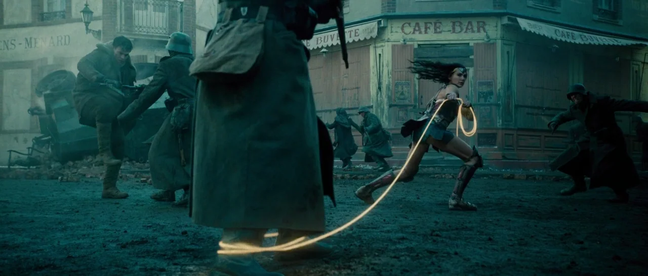 Wonder Woman (2017) movie still: wide — A war-torn village square with French-style cafe buildings and rubble on the ground. A woman in armor…