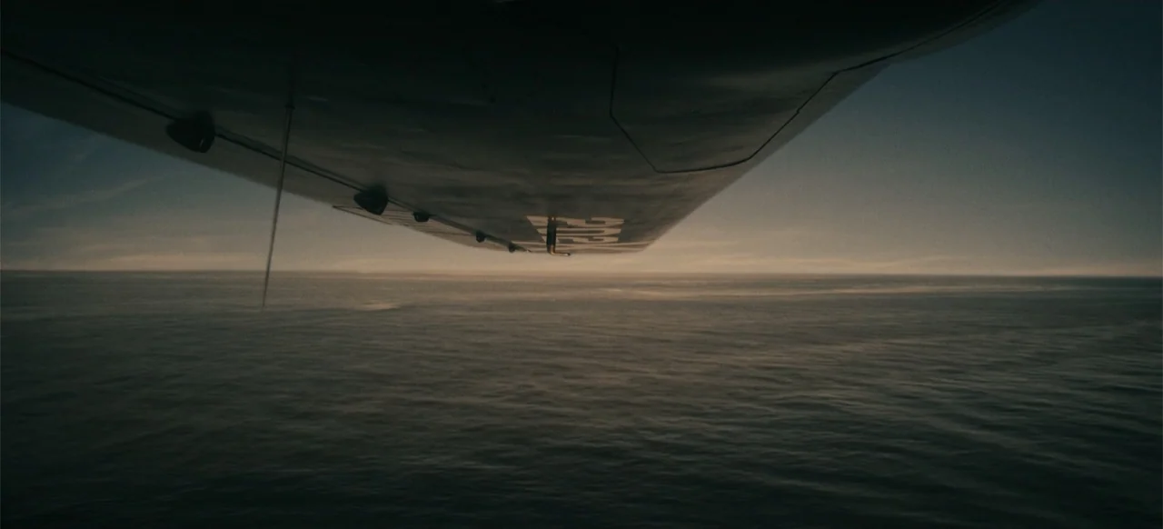 Devotion (2022) movie still: establishing — A dark, calm ocean stretches toward the horizon under a soft sunset sky. The underside of an airplane…