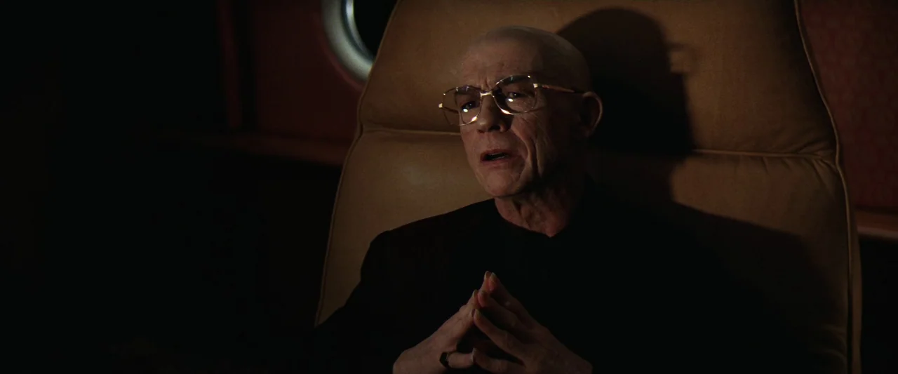Contact (1997) movie still: medium — An elderly man sits in a leather chair within a dim, enclosed cabin space. He holds his hands together while…