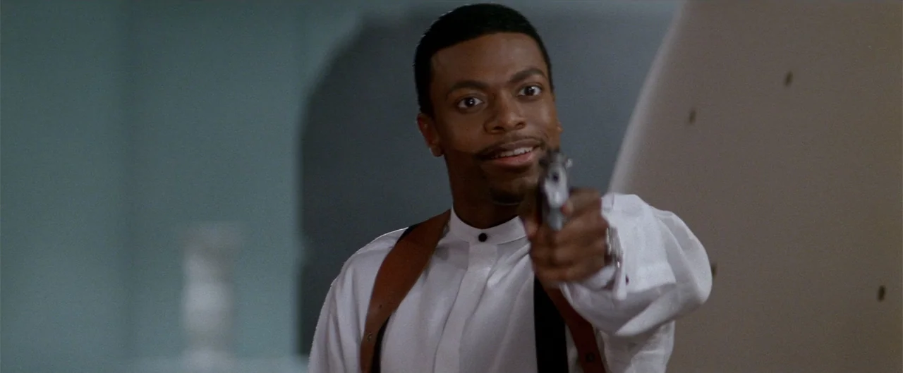Rush Hour (1998) movie still: medium — A dimly lit indoor space with plain walls and soft overhead lighting. A man in a white shirt holds a handgun…