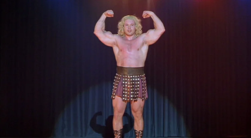 Invincible (2001) movie still: fullbody — A dimly lit stage features dark fabric curtains as a backdrop for a performer. A muscular man with blonde…