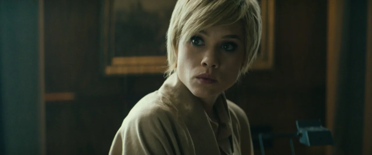 The Vault (2021) movie still: closeup — A dark, wood-paneled room interior with a framed picture visible in the background. A woman with short…