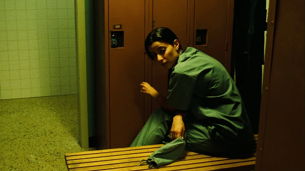 Saw III (2006) movie still: medium — A windowless locker room contains tall brown metal units and a wooden bench. A woman wearing green scrubs sits…