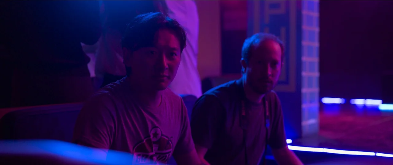 The Monk and The Gun (2023) movie still: medium — Two men sit in a dimly lit arcade interior saturated with colorful neon light. They both look…