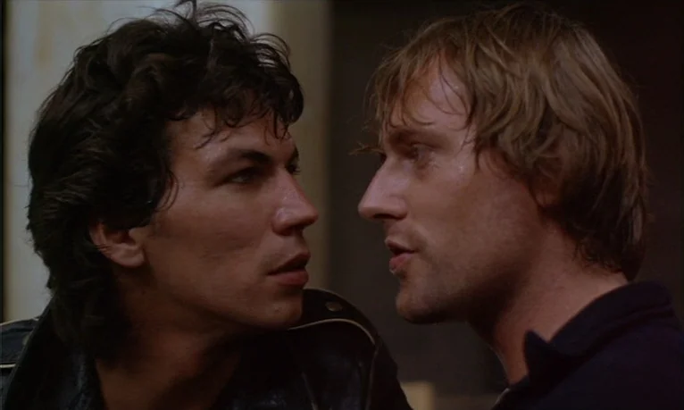 Spetters (1980) movie still: closeup — Two men stand in a dimly lit interior space with plain walls. One man in a leather jacket faces a man in a…