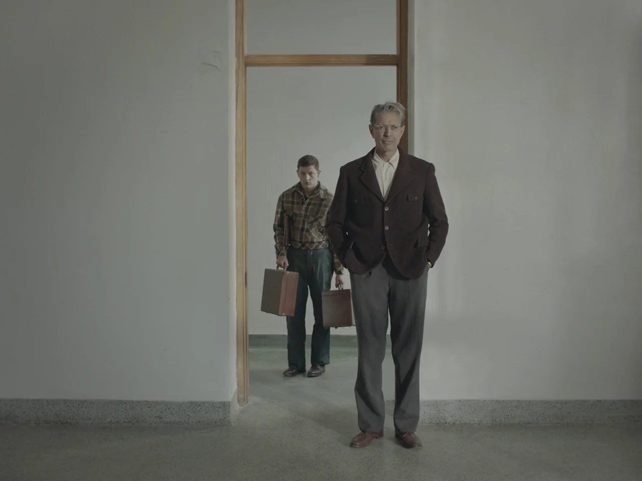 The Mountain (2018) movie still: fullbody — A stark, minimalist corridor with white walls and a light concrete floor. Two men stand in sequence…