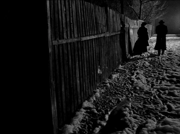 My 20th Century (1989) movie still: wide — A snowy, unpaved lane runs alongside a tall wooden picket fence at night. Two figures in long coats walk…