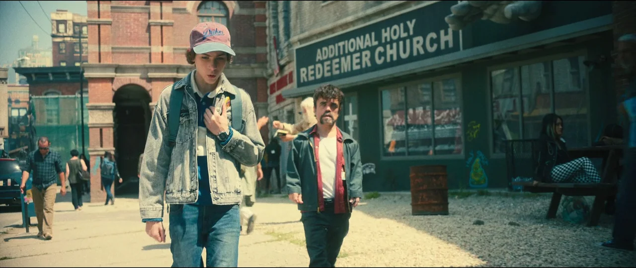 The Toxic Avenger (2023) movie still: fullbody — A city sidewalk fronting a church building with brick masonry and large glass windows. Two men walk…