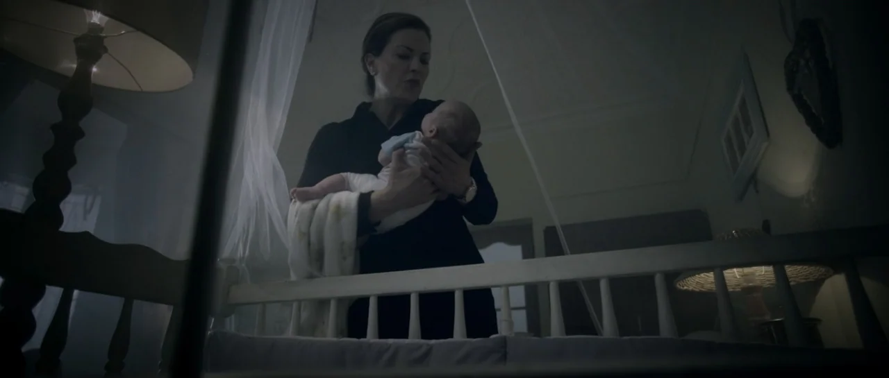 The Lullaby (2019) movie still: medium — A dark, dimly lit nursery features a white wooden crib and sheer white curtains. A woman holds an infant…