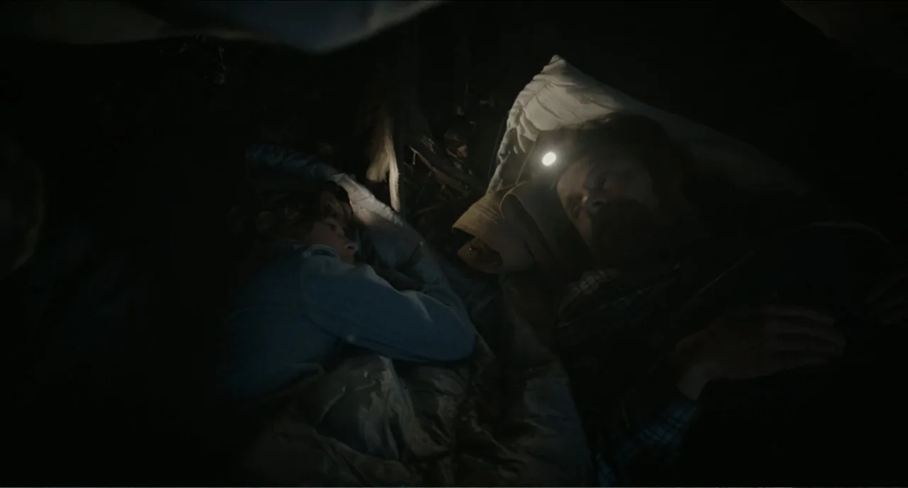 Cowboys (2014) movie still: medium — An adult man and a child are sleeping inside a dark tent together. The man wears an active headlamp that casts…