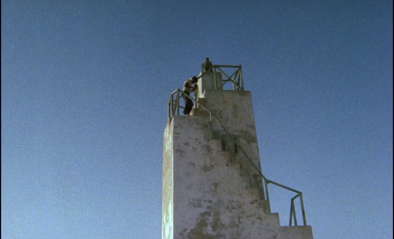 Sans Soleil (1983) movie still: wide — A weathered, stone watchtower stands against a vast, clear blue sky. A man walks up the exterior stairs…