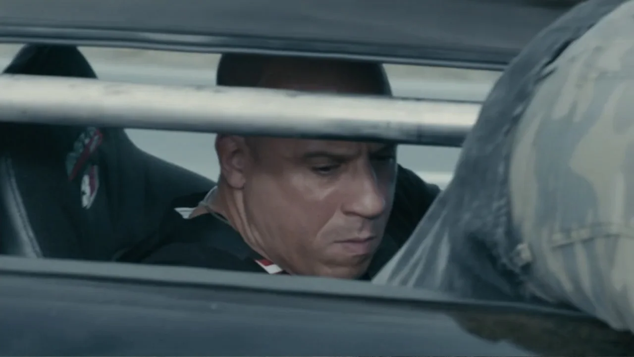 Furious 7 (2015) movie still: closeup — The interior of a vehicle shows a man sitting in the driver seat. A metal roll cage bar cuts horizontally…