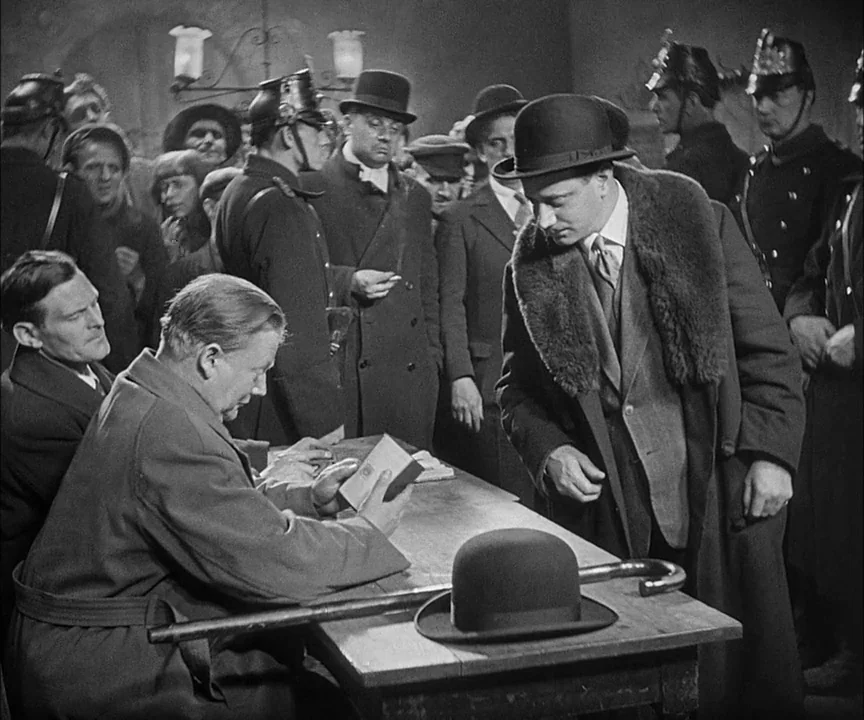 M (1931) movie still: medium — A crowded, dimly lit administrative room contains men in coats and period military helmets. A man sits at a wooden…