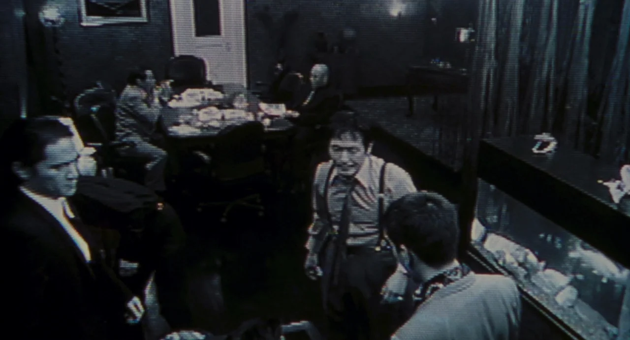 Ichi The Killer (2001) movie still: wide — A dimly lit room with a conference table is viewed from an overhead angle. Several men stand and sit in…