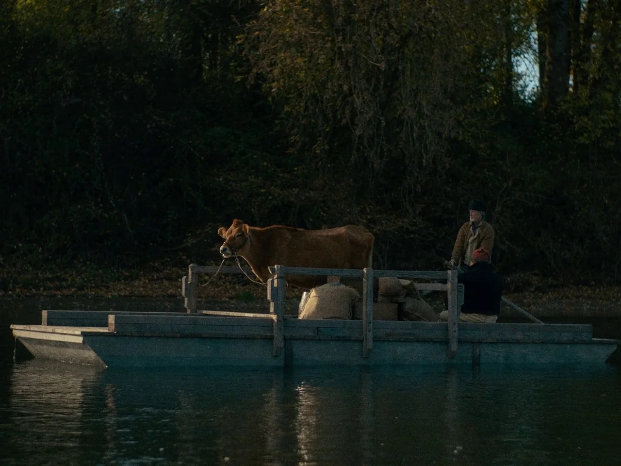 First Cow (2019) movie still: wide — A wooden barge floats on a dark river with a dense treeline in the background. Two men sit on the craft beside…