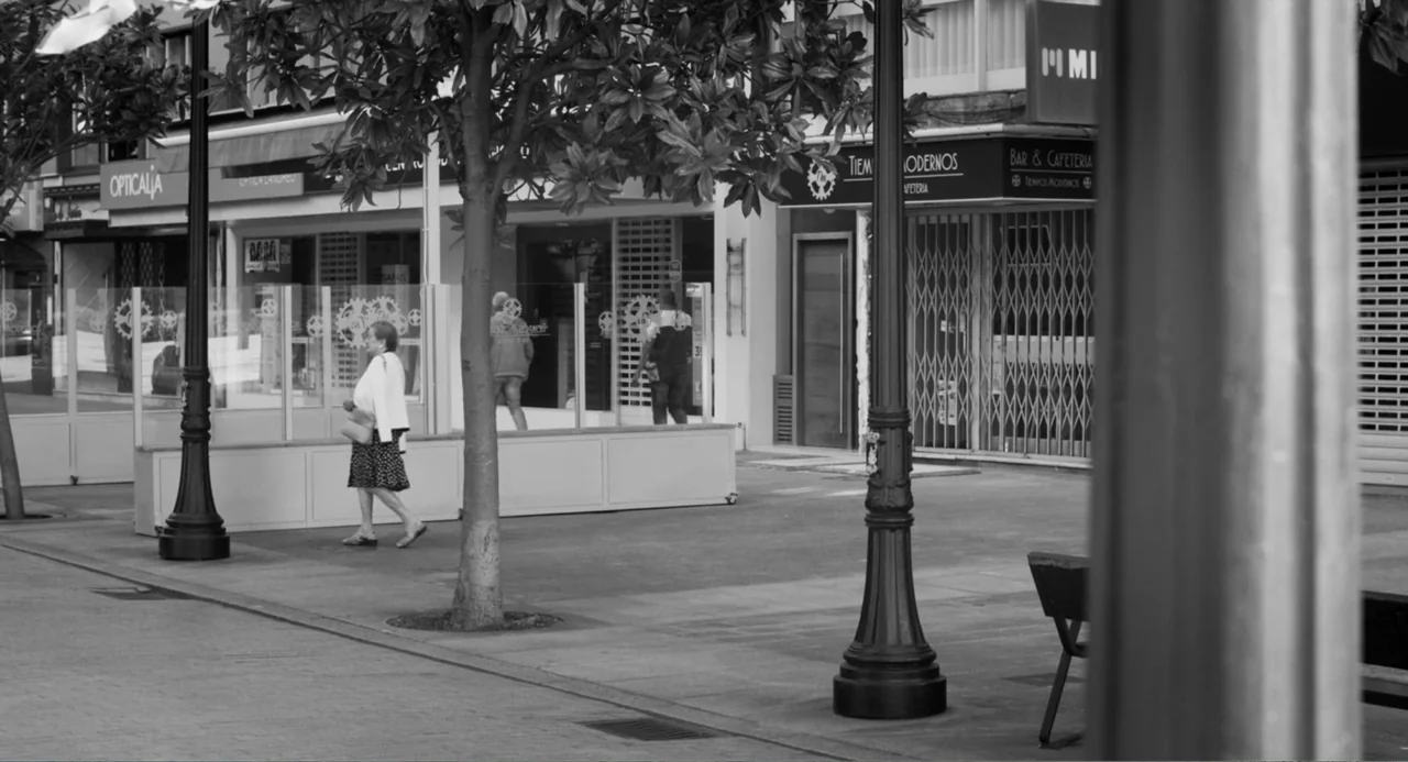 El Planeta (2021) movie still: fullbody — An empty urban sidewalk stretches past several glass-fronted shops with metal security shutters. An older…