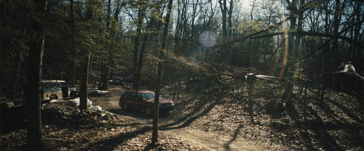 Skin (2009) movie still: wide — A dense forest with bare trees surrounds a dirt path scattered with dry leaves. Two vehicles are parked along the…