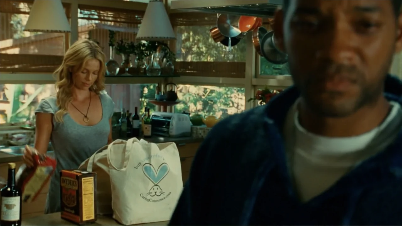 Hancock (2008) movie still: medium — A sunlit residential space with glass-fronted cabinets and natural wood finishes. A man stands in the blurred…