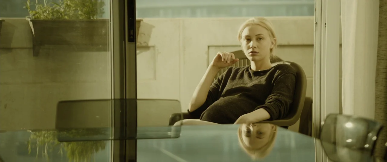 Enemy (2013) movie still: medium — A woman sits on a chair on a balcony overlooking a bright area. The subject rests in the sunlight with a glass…